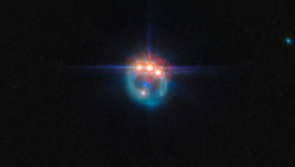 Una piccola immagine di una galassia distorta dalla lente gravitazionale in un anello fioco. Nella parte superiore dell'anello ci sono tre punti molto luminosi con punte di diffrazione che fuoriescono da essi, uno accanto all'altro: in realtà si tratta di copie di un singolo quasar nella galassia, duplicate dalla lente gravitazionale. Al centro dell'anello, la galassia ellittica che esegue la lente appare come un piccolo punto blu.